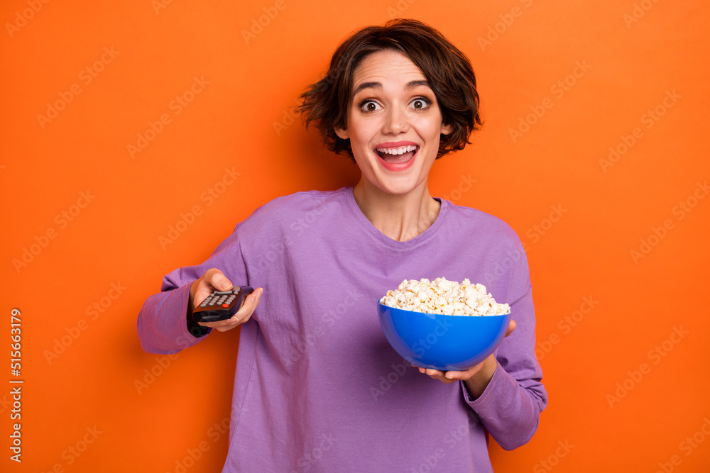 Photo of astonished positive lady hold popcorn plate remote control switch channel isolated on orange color background Photo of astonished positive lady hold popcorn plate remote control switch channel isolated on orange color background