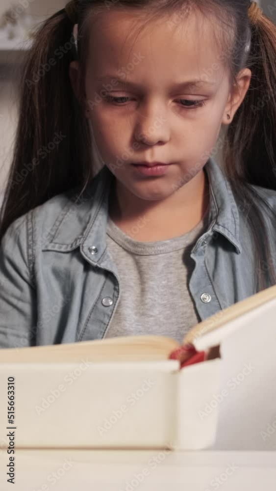 Vertical video. Kid literature. Schoolgirl study. Intelligent child hobby. Tired cute smart small girl reading book at home workplace. Vertical video. Kid literature. Schoolgirl study. Intelligent child hobby. Tired cute smart small girl reading book at home workplace.