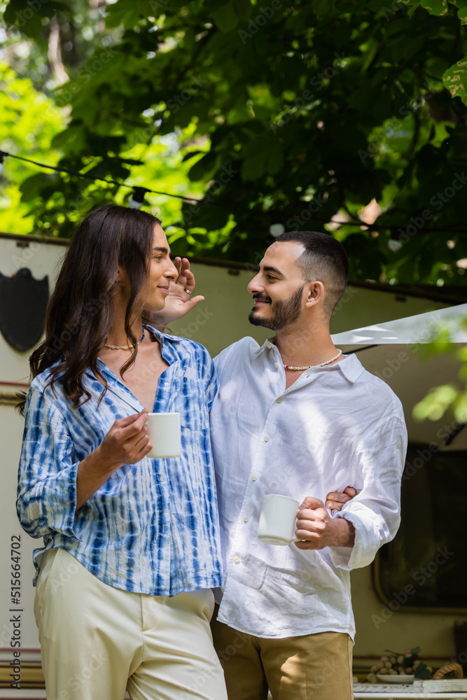 happy gay couple holding cups of coffee while looking at each other near van during journey in summer. happy gay couple holding cups of coffee while looking at each other near van during journey in summer.