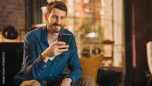 Portrait of a Handsome Man Sitting on Couch, Using Smartphone in Stylish Loft Apartment or Cafe. Creative Male Smiling, Checking Social Media and Typing Message. Urban City View from Big Window.