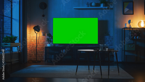 Stylish Loft Apartment Interior with TV Set with Green Screen Mock Up Display Standing on Television Stand. Empty Living Room at Home with Chroma Key Placeholder on Monitor. Night Shot. photo