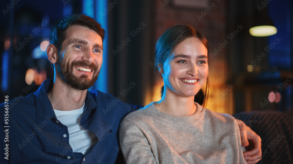 Portrait of Beautiful Couple Spending Time at Home, Sitting on a Couch, Hugging and Watching Exciting TV Show in Their Stylish Loft Apartment. Man and Woman Streaming Comedy Movie and Have Good Time. Portrait of Beautiful Couple Spending Time at Home, Sitting on a Couch, Hugging and Watching Exciting TV Show in Their Stylish Loft Apartment. Man and Woman Streaming Comedy Movie and Have Good Time.