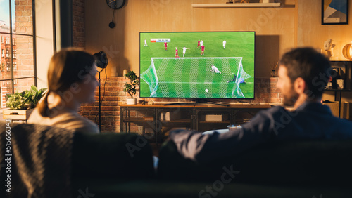 Couple of Soccer Fans Relax on a Couch, Watch a Sports Match at Home in Stylish Loft Apartment. Relaxed young Man and Woman Cheer for Their Favorite Football Club and Enjoying the Evening. photo