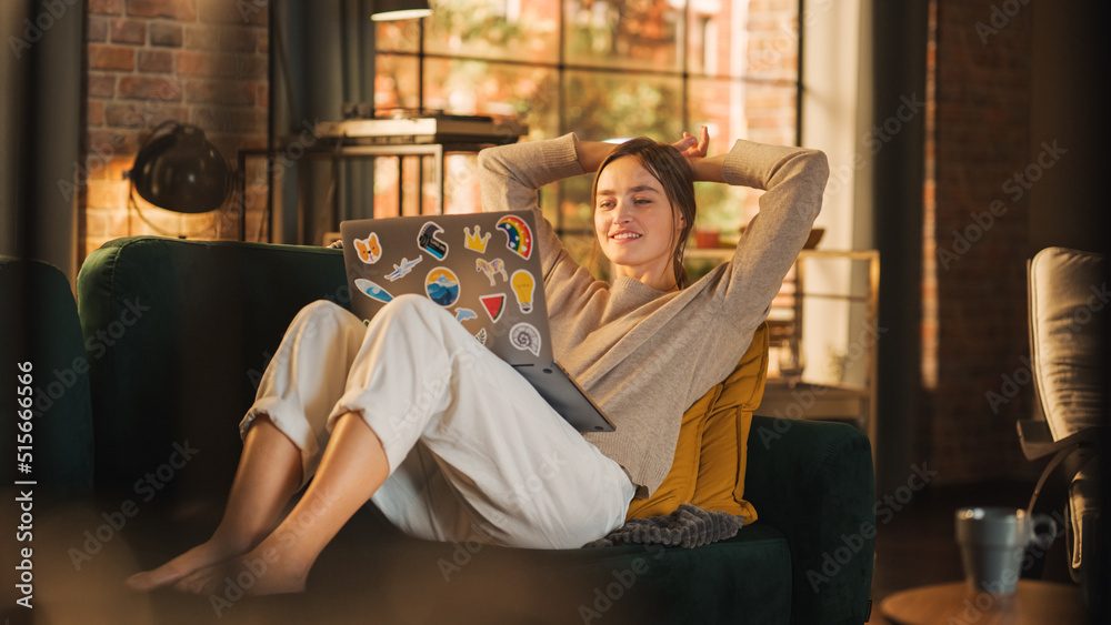 Young Beautiful Woman Lying on Couch, Using Laptop Computer in Stylish Loft Apartment on a Sunny Day. Creative Female Smiling, Checking Social Media, Typing Message. Urban City View from Big Window. Young Beautiful Woman Lying on Couch, Using Laptop Computer in Stylish Loft Apartment on a Sunny Day. Creative Female Smiling, Checking Social Media, Typing Message. Urban City View from Big Window.