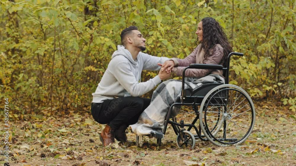 Young happy hispanic couple in love having cute conversation talking caring boyfriend visits sick girlfriend in hospital park woman patient in wheelchair needs rehabilitation loving guy supports girl Young happy hispanic couple in love having cute conversation talking caring boyfriend visits sick girlfriend in hospital park woman patient in wheelchair needs rehabilitation loving guy supports girl