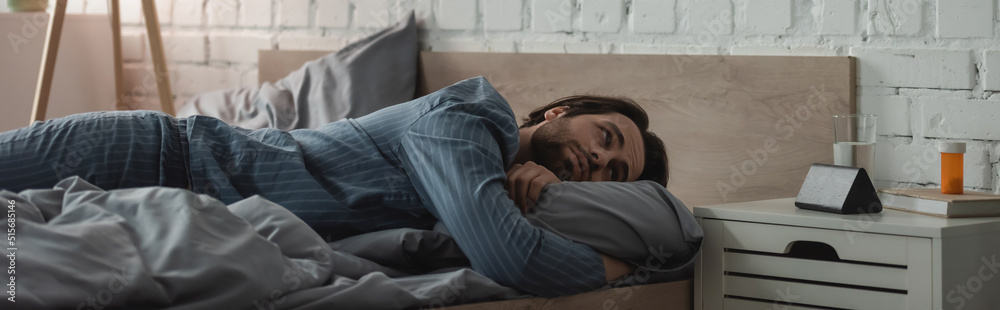Bearded man looking at alarm clock near water and pills on bedside table in bedroom, banner Bearded man looking at alarm clock near water and pills on bedside table in bedroom, banner