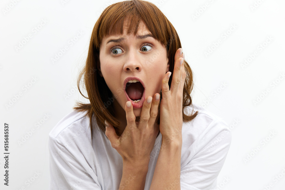 a frightened, shocked, emotional woman screams loudly clutching her face and with very frightened, wide-open eyes stands on a light background in a white T-shirt. a frightened, shocked, emotional woman screams loudly clutching her face and with very frightened, wide-open eyes stands on a light background in a white T-shirt.