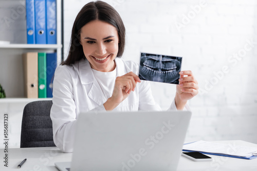 Positive doctor holding scan of teeth during video chat on laptop in hospital