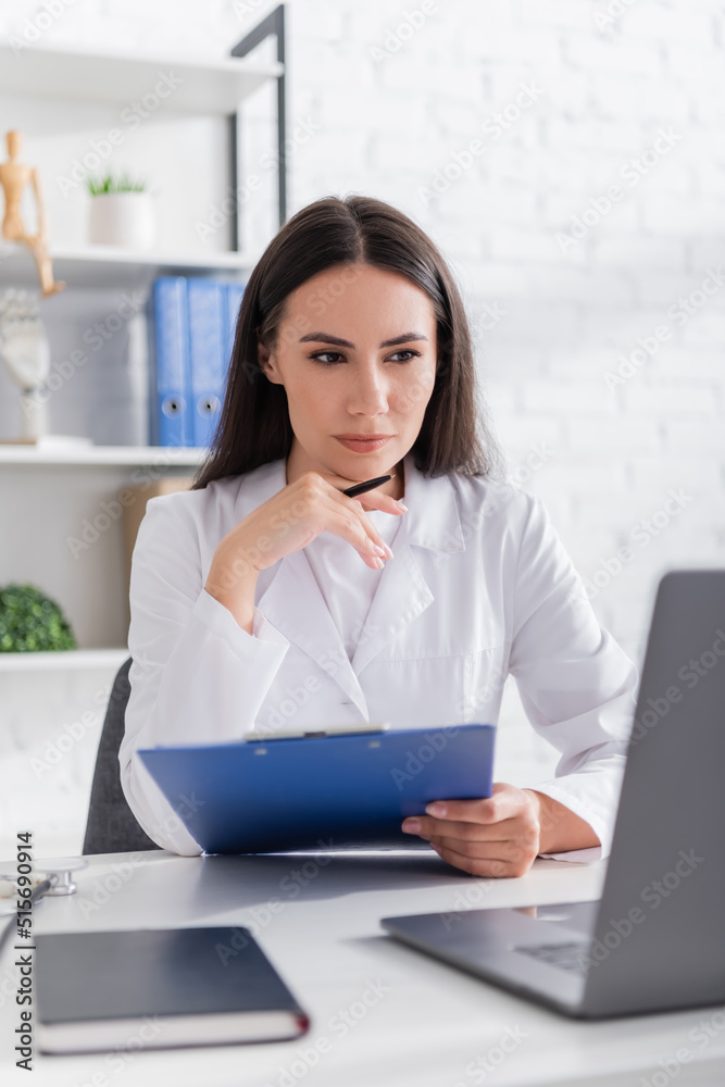 Doctor holding pen and clipboard and looking at blurred laptop in clinic Doctor holding pen and clipboard and looking at blurred laptop in clinic