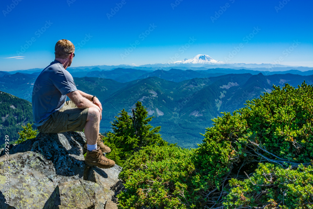 An athletic adventurous male hiker sitting on top of a mountain looking out at a mountain range on a sunny day.