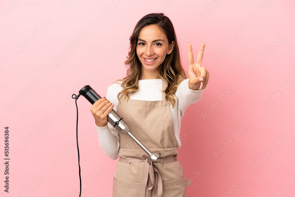Young woman using hand blender over isolated pink background smiling and showing victory sign Young woman using hand blender over isolated pink background smiling and showing victory sign