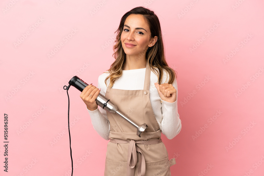 Young woman using hand blender over isolated pink background proud and self-satisfied