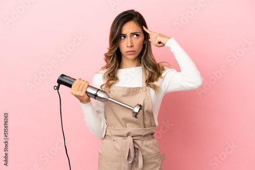 Young woman using hand blender over isolated pink background having doubts and thinking