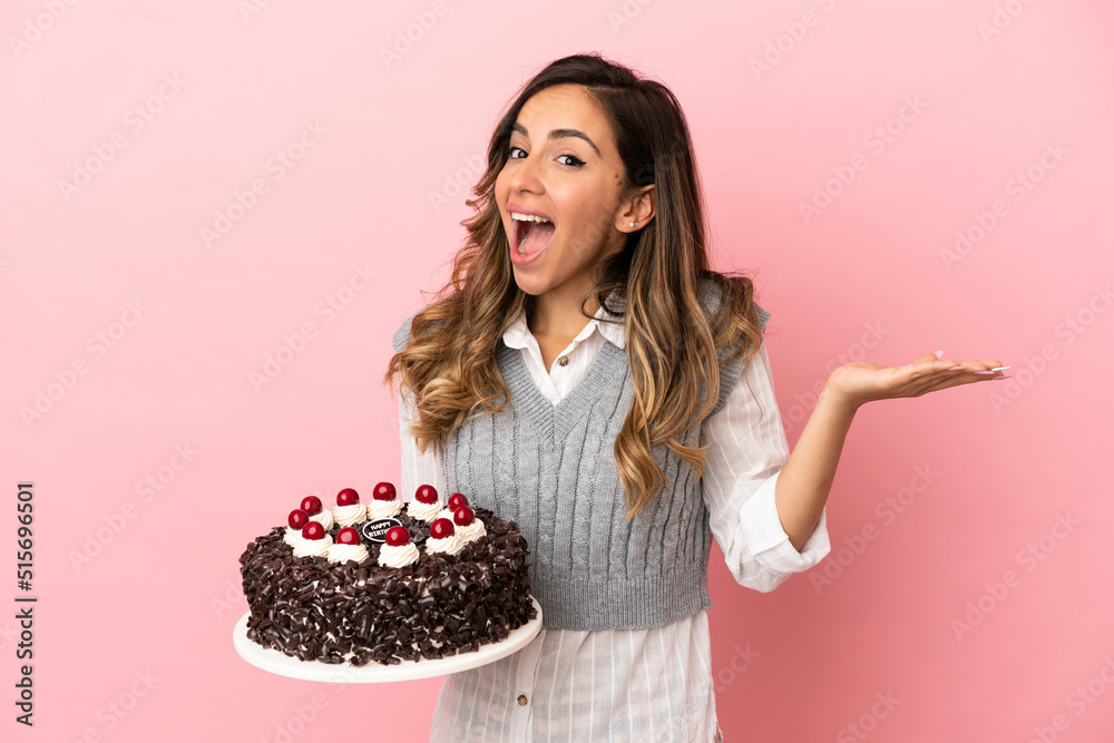Young woman holding birthday cake over isolated pink background with shocked facial expression Young woman holding birthday cake over isolated pink background with shocked facial expression