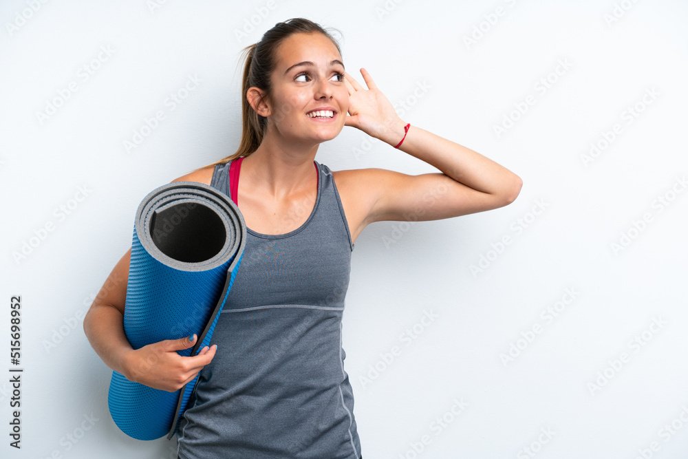 Young sport woman going to yoga classes while holding a mat isolated on white background listening to something by putting hand on the ear Young sport woman going to yoga classes while holding a mat isolated on white background listening to something by putting hand on the ear