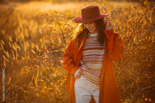 Happy smiling fashionable curly brunette woman wearing trendy autumn orange trench coat, patterned sweater, white jeans, stylish hat, posing in autumn nature during sunset. Copy, empty space for text  © Victoria Fox