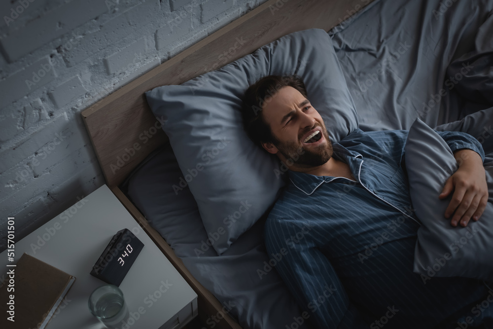 Top view of man with insomnia yawning near clock and water on bedside table in morning Top view of man with insomnia yawning near clock and water on bedside table in morning