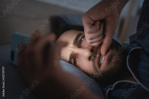 Stressed man biting nail on hand and using smartphone on bed at night