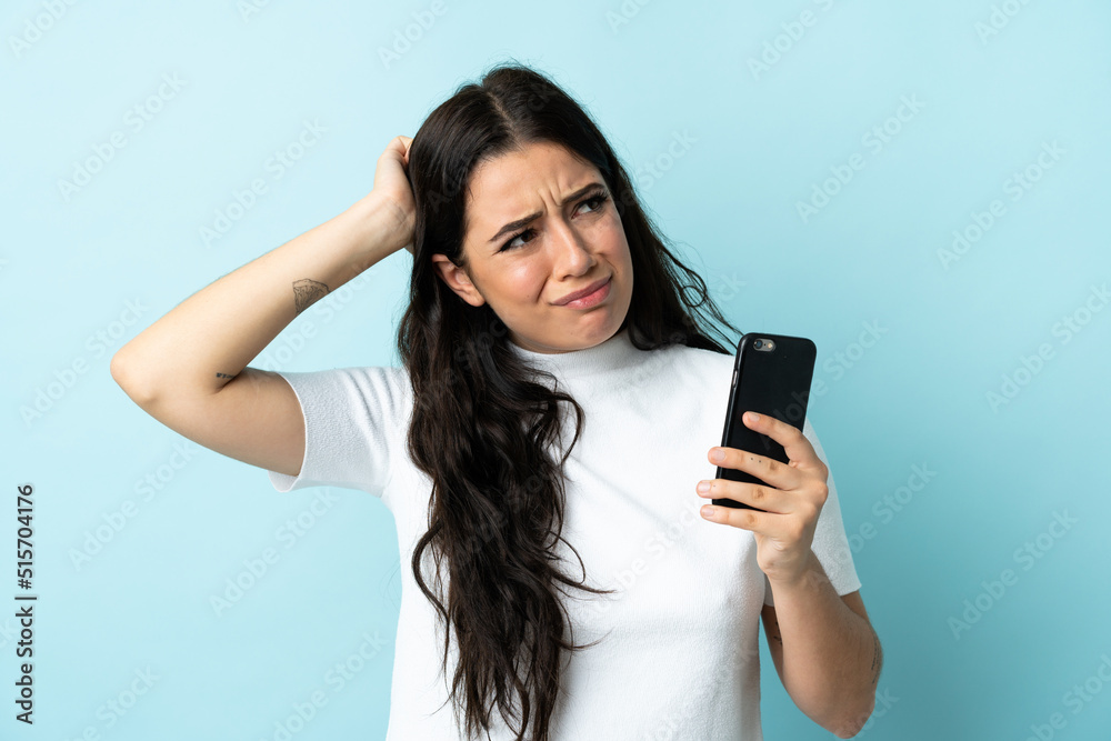 Young woman using mobile phone isolated on blue background having doubts and with confuse face expression Young woman using mobile phone isolated on blue background having doubts and with confuse face expression