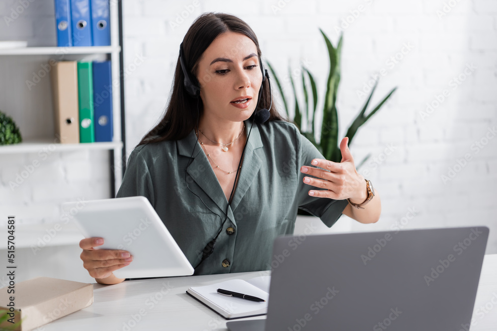 Tutor in headset holding digital tablet during online lesson on laptop in school