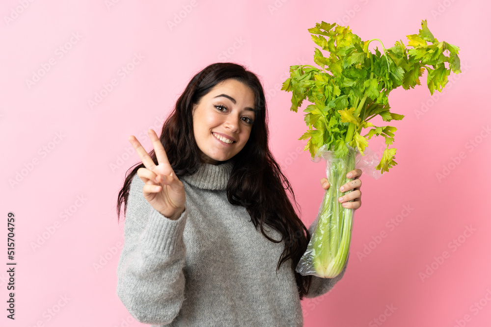Young caucasian woman holding a celery isolated on blue background smiling and showing victory sign Young caucasian woman holding a celery isolated on blue background smiling and showing victory sign