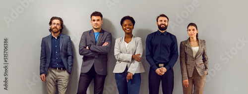 Confident business team in modern smart clothes. Group of happy young diverse people in formal suits standing in fashion studio and leaning on gray wall. Office dress code concept. Banner  header