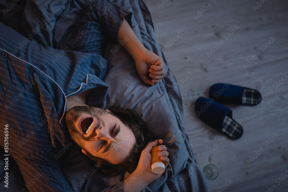 Top view of man with insomnia holding pills and yawning on bed