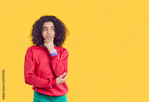 Cute smiling teen girl dreams, thinks, makes decisions or comes up with cunning plan. African American curly girl with sly expression looks at copy space on yellow background. Web banner.
