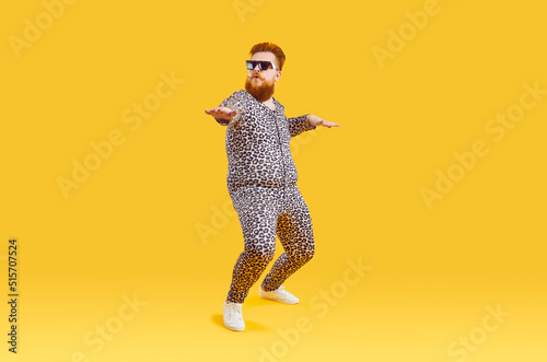 Plus size male model in funny PJs having fun in modern fashion studio. Happy carefree confident fat bearded man in comfortable leopard pajamas and cool glasses dancing isolated on yellow background photo