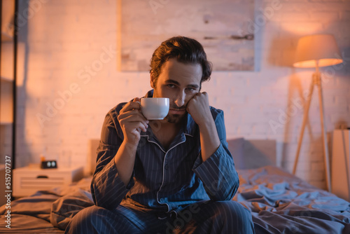 Obraz na plátně Displeased man with sleep disorder holding cup and looking at camera on bed