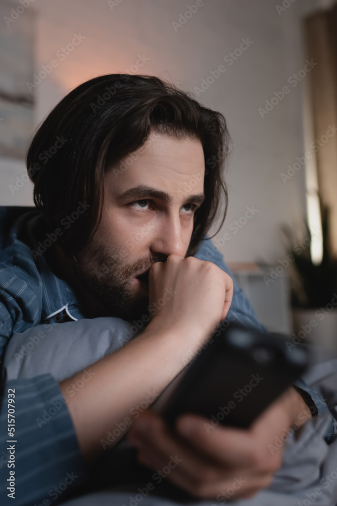 Fototapeta premium Stressed man with insomnia holding blurred remote controller on bed at night