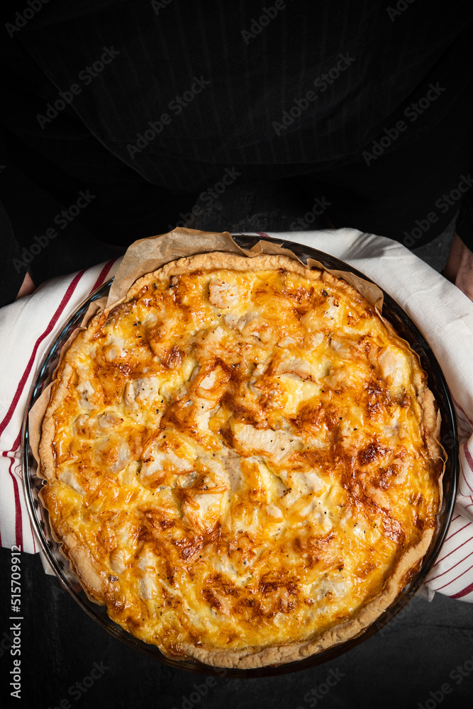 Quiche pie in chef hands on dark background. Savory tart Lorraine with cheese, chicken and mushrooms, freshly baked in oven. Traditional french cuisine. Home cooking recipe. Vertical, copy space