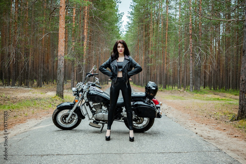 A girl in tight black leather stands next to a motorcycle.