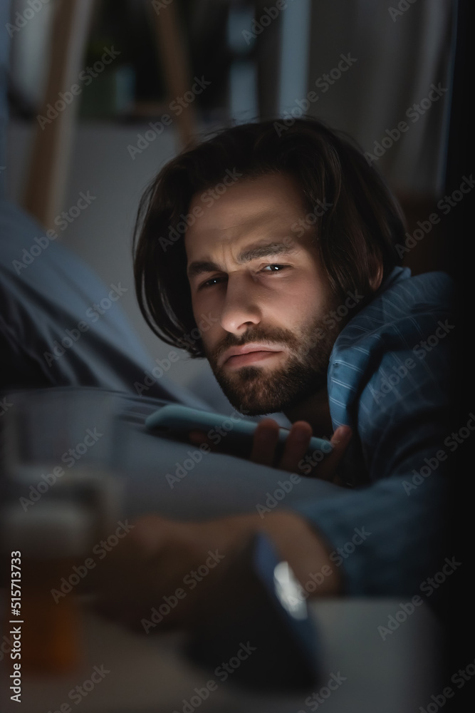 Obraz premium Dissatisfied man with insomnia holding smartphone on bed at home