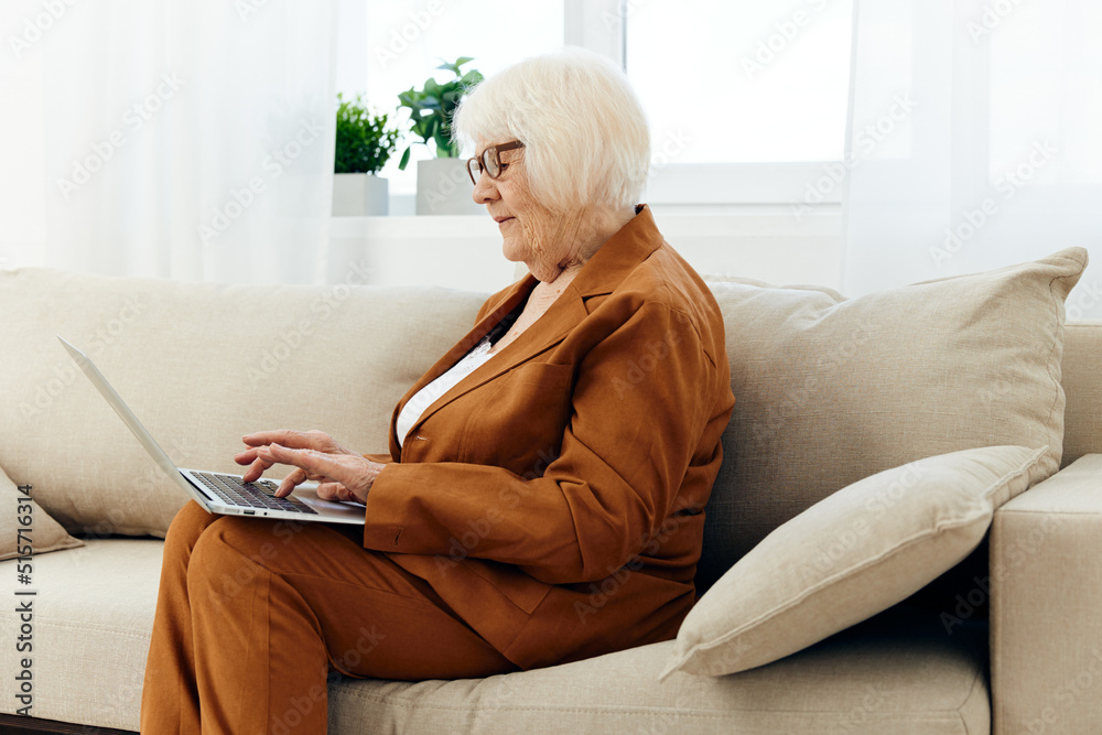 an elderly businesswoman is sitting on a beige sofa in her bright apartment and carefully looks at her laptop while working remotely behind it an elderly businesswoman is sitting on a beige sofa in her bright apartment and carefully looks at her laptop while working remotely behind it