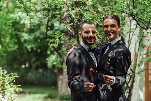 happy gay couple in formal wear holding sparklers in green park on wedding day.