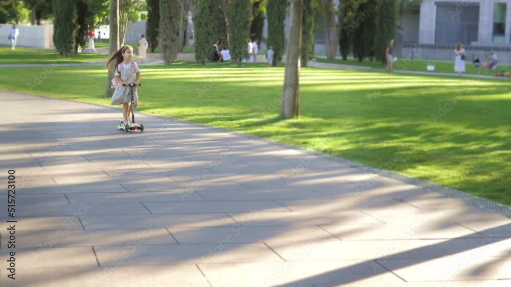 A little girl in a dress rides through the park on her scooter. Happy childhood, lifestyle. A Caucasian child rides a scooter on a path on a bright sunny day. Children's games, entertainment A little girl in a dress rides through the park on her scooter. Happy childhood, lifestyle. A Caucasian child rides a scooter on a path on a bright sunny day. Children's games, entertainment