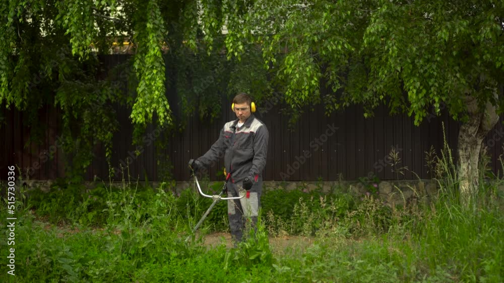 A young man from the special services mows the lawn with a petrol trimmer. A man in glasses and headphones with a lawn mower in his hands. A young man from the special services mows the lawn with a petrol trimmer. A man in glasses and headphones with a lawn mower in his hands.