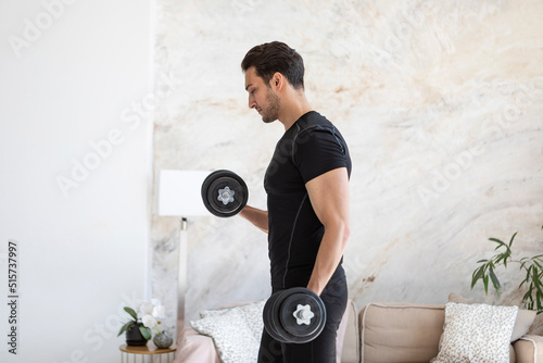 Side view of young handsome man in black sportswear doing biceps curl with dumbbells in living room at home. Healthy lifestyle and home workout concept