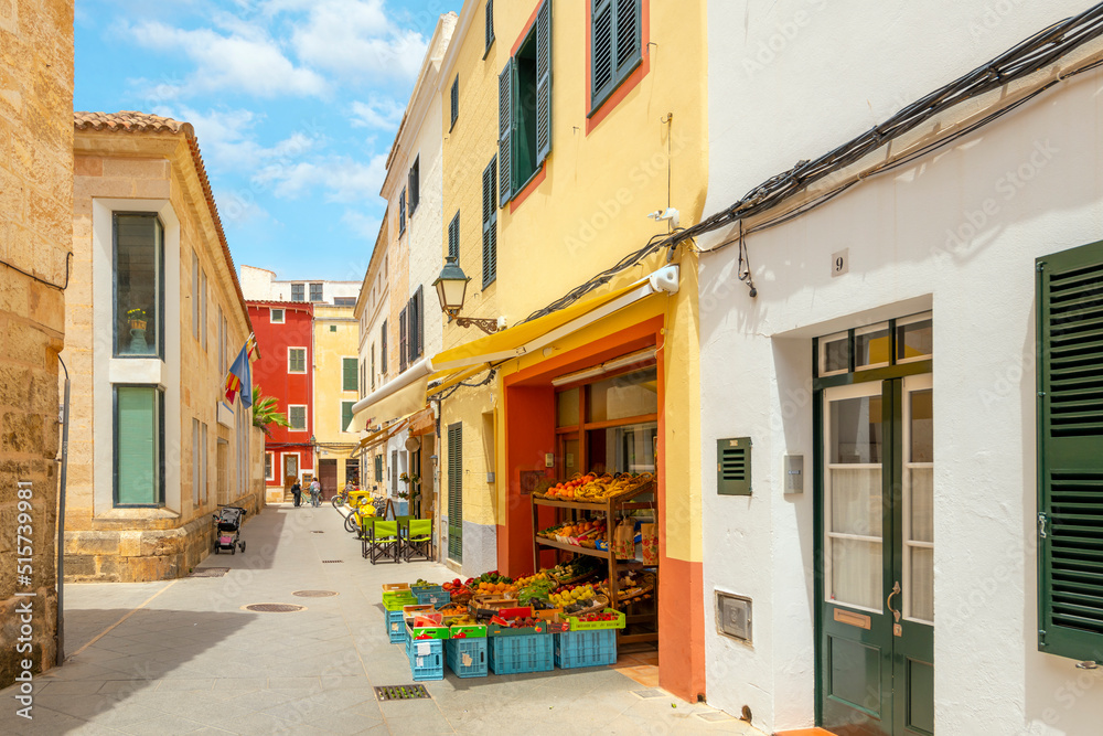 A narrow street of shops including a produce market with fruit and vegetables on display in the historic Spanish town of Ciutadella de Menorca.