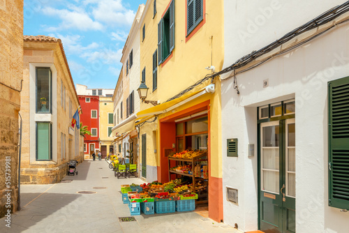 A narrow street of shops including a produce market with fruit and vegetables on display in the historic Spanish town of Ciutadella de Menorca.
