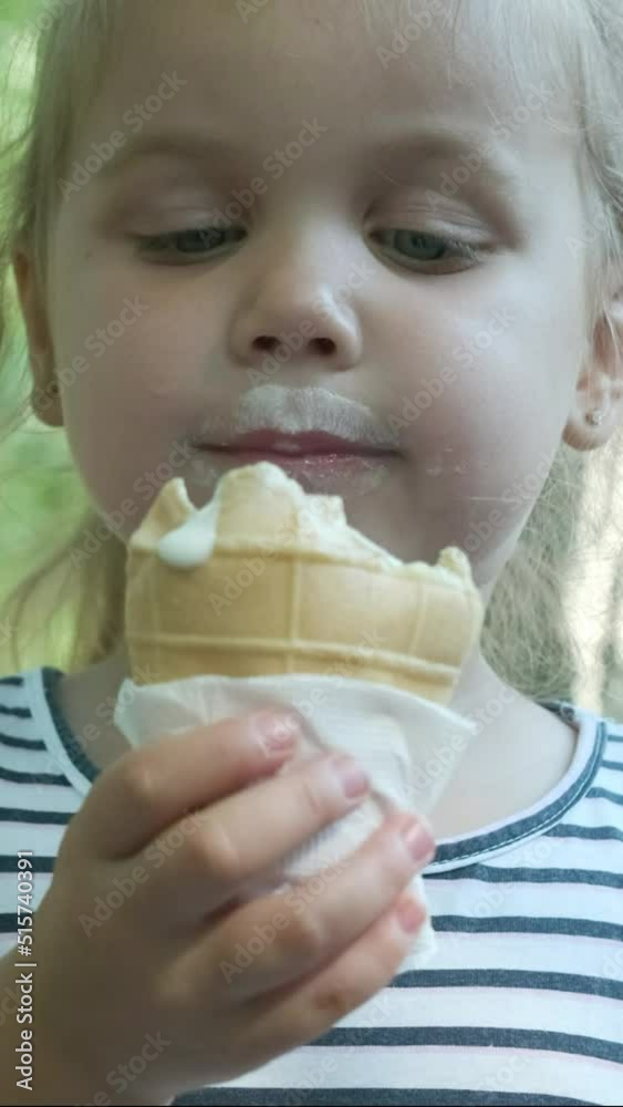 VERTICAL VIDEO: Smiling little girl eats ice cream. Close-up portrait of blonde girl sitting on park bench and eating icecream and smiles. Slow motion, Camera slowly moving forwards VERTICAL VIDEO: Smiling little girl eats ice cream. Close-up portrait of blonde girl sitting on park bench and eating icecream and smiles. Slow motion, Camera slowly moving forwards
