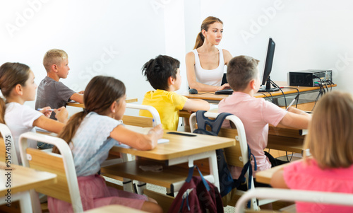 Positive female teacher working with computer and giving lesson for primary school pupils in classroom
