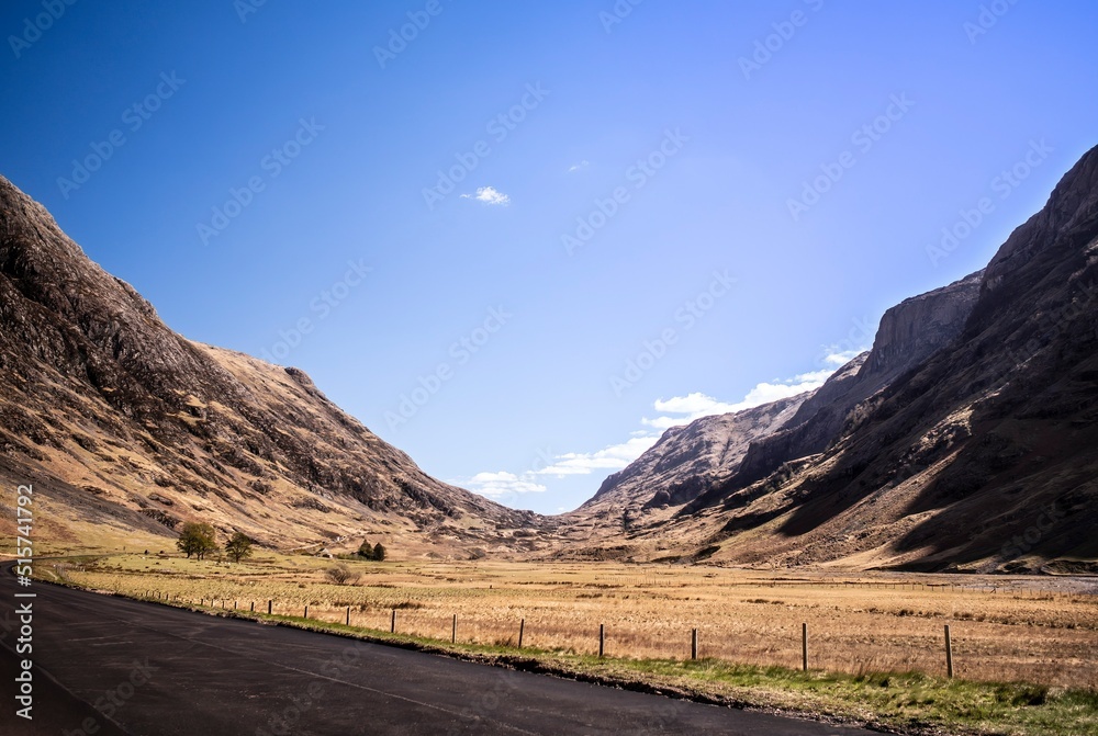 Road curving next to brown, grassy field in the Scottish Highlands near An Torr and Glencoe. Clear blue sky above, mountains on either side. Scotland, UK. Road curving next to brown, grassy field in the Scottish Highlands near An Torr and Glencoe. Clear blue sky above, mountains on either side. Scotland, UK.