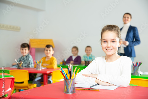 Cute intelligent girl sitting at school desk in classroom on background with classmates and teacher