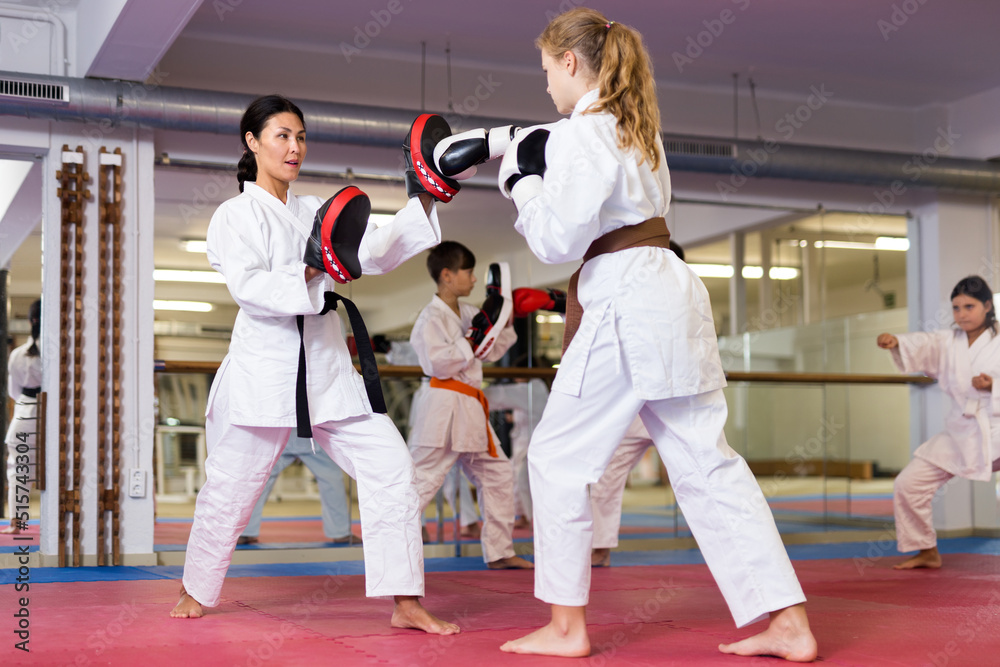 Young girls and boys in boxing gloves exercising jabs during group karate training. Woman trainer with focus mitts teaching girl in foreground.