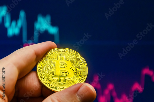 Golden coin of bitcoin in a man hand on the background of the screen with a chart of the bitcoin rate. Close up photo