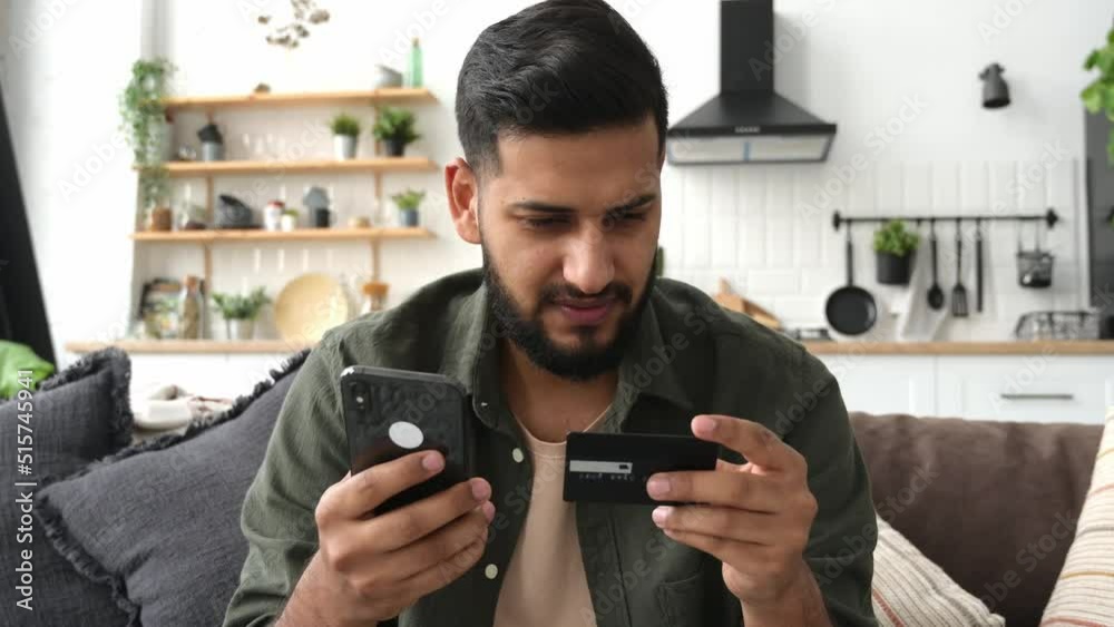 Online shopping, payment. Satisfied Indian or Arabic attractive guy, sit on the sofa in the living room, using a bank card and a smartphone, making online purchases, making payments, ordering delivery