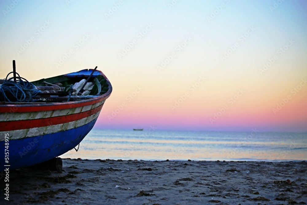 Fototapeta premium Boat with faded multi-color paint standing on the beach sand. In the background pink and blue light of dawn over the sea.
