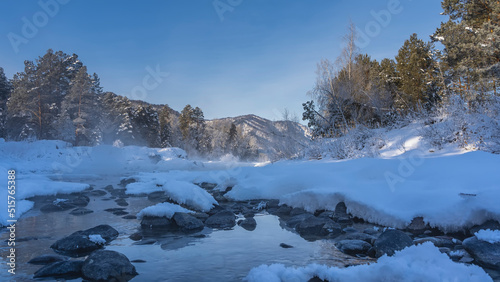 The ice-free river flows between the snow-covered banks. Steam over the water, hoarfrost on the rocks. Coniferous forest and mountains against the blue sky. Altai